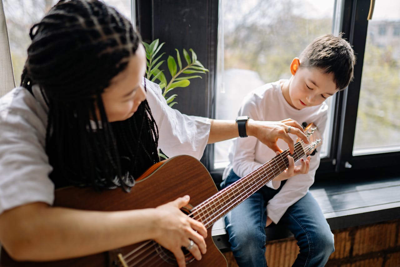 A child learns guitar with an adult by a window, enjoying a sunny day indoors.