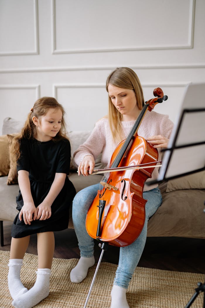 A young girl learns to play the cello at home with a female teacher, sitting together.